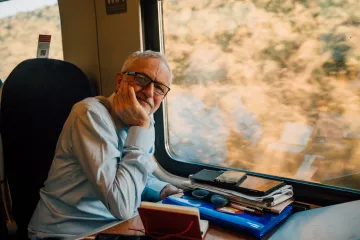 L'image montre un homme assis à côté d'une fenêtre de train. Il porte une chemise claire et a des lunettes. Son visage exprime une certaine réflexion ou contemplation alors qu'il repose sa tête sur sa main. Sur la table devant lui, on peut voir plusieurs objets comme des téléphones, des lunettes de soleil et des papiers, donnant l'impression qu'il a passé du temps à travailler ou à organiser ses affaires. À l'extérieur de la fenêtre, on aperçoit un paysage flou, suggérant que le train est en mouvement.