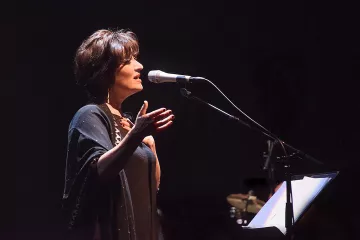 L'image montre une femme sur scène, en train de chanter ou de parler dans un microphone. Elle a les cheveux courts et bruns, et porte une tenue sombre, accompagnée d'un châle. Son expression semble engagée et passionnée, suggérant qu'elle interprète une performance émotionnelle. En arrière-plan, on aperçoit des instruments de musique, ce qui indique qu'elle se produit dans un cadre musical. L'éclairage est tamisé, créant une ambiance intime et captivante.