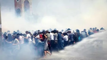 L'image montre une scène de manifestation où un grand groupe de personnes semble être en mouvement, probablement en train d'échapper à des gaz lacrymogènes ou à une intervention des forces de l'ordre. Le brouillard blanc, causé par la fumée ou les sprays, rend l'atmosphère tendue et chaotique. En arrière-plan, une structure architecturale est visible, mais elle est à peine discernable à cause de la fumée. Les manifestants semblent rassemblés et stressés, certains essayant de se protéger ou de se diriger vers une zone plus sûre.