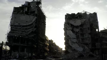 تظهر الصورة عمارتين محطمتين في منطقة يبدو أنها تعرضت لتدمير واسع. المباني مشوهة، حيث تظهر الجدران المنهارة والأسطح المدمرة، مما يعكس آثار الدمار. السماء في الخلفية تبدو ملبدة، مما يضيف شعورًا بالخيبة والخراب. المنطقة المحيطة يبدو أنها مهجورة، مما يعكس الأثر العميق للصراعات أو الكوارث التي أدت إلى هذا الوضع.