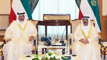 L'image montre deux hommes assis dans une salle de réunion élégante. Ils portent des vêtements traditionnels, possiblement des habitudes de type arabe, avec des écharpes sur la tête. Leurs expressions sont sérieuses et ils sont entourés de décorations luxueuses, comme des rideaux drapés et des meubles en bois. Au centre, il y a une table en marbre avec quelques objets, et en arrière-plan, on peut voir un tableau avec des inscriptions arabes. L'atmosphère dégage un sentiment de formalité et de respect.