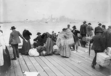 L'image montre un groupe de personnes rassemblées sur un quai, probablement à une époque antérieure. On peut voir des individus portant des habits typiques de l'époque, dont des chapeaux, et certains sont emmitouflés dans des couvertures. Ils semblent s'affairer autour de bagages et de marchandises, évoquant des thèmes de migration ou de transit. En arrière-plan, on aperçoit des bâtiments ainsi que des bateaux sur l'eau, suggérant un port animé. L'atmosphère semble à la fois chargée et historique.