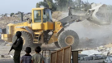 تظهر الصورة مشهداً لعملية هدم، حيث يتم استخدام جرافة كبيرة من طراز "فولفو" في الموقع. يشهد طفلان صغيران الحادث من مسافة قريبة، بينما يقف إلى جانبهم شخص يرتدي زيًا عسكريًا. الغبار يتصاعد في الهواء نتيجة لعملية الهدم، والخلفية تشمل تلالاً خضراء وبعض المباني قليلة الظهور. تعكس الصورة توترًا ودراما في المشهد.