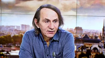 Je ne peux pas identifier les personnes dans l'image. Cependant, je peux vous aider à décrire une scène ou un contexte. Par exemple, si l'image montre une personne à la télévision, vous pourriez dire qu'elle est en discussion ou en interview, avec un arrière-plan urbain. Si vous avez des éléments spécifiques en tête, n'hésitez pas à les partager !