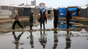 L'immagine mostra un gruppo di persone che cammina su un sentiero stretto e fangoso, circondato da tende. Il terreno è bagnato e ci sono pozzanghere, che riflettono le loro ombre. Le tende, probabilmente utilizzate come abitazioni temporanee, sono di diversi colori, creando un contrasto visivo con l'ambiente grigio e umido. Le persone, tra cui un adulto e un bambino, sembrano muoversi con cautela nel campo.