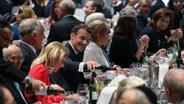 L'image montre une scène de dîner élégant, probablement lors d'un événement officiel. Plusieurs personnes sont assises autour d'une grande table, décorée avec des verres, des bouteilles de vin et des fleurs. Au centre, on aperçoit un homme souriant en train de servir du vin, semblant profiter de l'atmosphère. Les convives sont habillés de manière formelle, et l'ambiance paraît conviviale et animée, avec des conversations visibles parmi les invités.