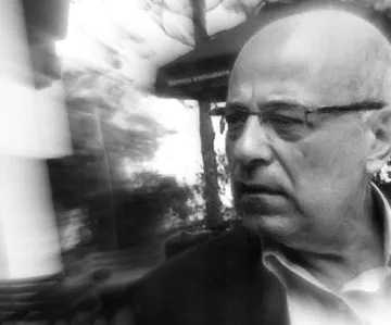 L'image présente un homme d'âge mûr avec des lunettes, légèrement flou en raison d'un effet de mouvement. Il semble pensif ou préoccupé, et son expression faciale transmet une certaine intensité. L'arrière-plan est indistinct, ce qui ajoute à l'effet dynamique de la photo. Les tonalités en noir et blanc renforcent le caractère dramatique de l'image.