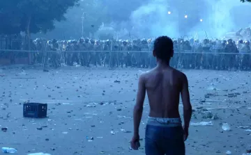 L'image montre un jeune garçon, torse nu, qui se tient de dos face à une ligne de forces de l'ordre en uniforme. Il est dans un environnement troublé, avec des débris et des bouteilles éparpillés au sol, ainsi qu'une légère brume qui donne une atmosphère tendue et chaotique. La scène semble se dérouler dans un contexte de troubles ou de manifestations. Les policiers, en arrière-plan, se tiennent en formation, suggérant un affrontement imminent.