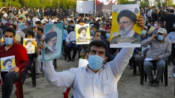 تظهر الصورة تجمعًا حاشدًا لمجموعة من الأشخاص يجلسون في مكان مفتوح. يحمل بعضهم لافتات أو صورًا لأحد الشخصيات السياسية. يرتدى الجميع تقريبًا كمامات واقية، مما يدل على الاحتراز من الأمراض. يبدو أن التجمع مرتبط بحدث سياسي أو دعم لشخص معين. الخلفية تضم أشجارًا ومباني، مما يعطي انطباعًا عن حدث في منطقة حضرية.