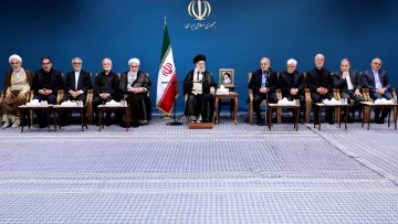 L'image montre un groupe de personnes assises en rangée sur un tapis, dans un environnement formel. En arrière-plan, il y a un drapeau iranien et un portrait d'une figure importante, probablement un leader. Les participants semblent être des hommes en costume sombre et en tenue religieuse, certains portant des turbans. Ils semblent engagés dans une discussion ou une réunion officielle. L'ambiance est solennelle et professionnelle.
