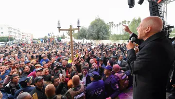 L'image montre un homme, probablement un orateur, s'adressant à une grande foule de personnes. La scène semble se dérouler à l'extérieur, avec des arbres en arrière-plan. Le public est très nombreux et semble attentif à ce qui est dit. À l'avant de la scène, des membres des forces de sécurité ou des agents en uniforme sont présents pour assurer l'ordre. L'atmosphère paraît dynamique et engagée, suggérant un événement public important.