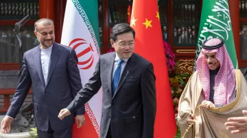 تظهر في الصورة ثلاثة رجال يقفون أمام أعلام ثلاث دول: إيران، الصين، والسعودية. الشخص في الوسط يبدو أنه يتحدث أو يتحرك، بينما الشخصان على الجانبين ينظران إليه. الأجواء تبدو رسمية، وقد تكون الصورة ملتقطة خلال حدث دبلوماسي أو اجتماع. الخلفية تحتوي على نباتات وزينة أخرى تضفي طابعاً احتفائياً.