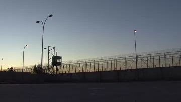 L'image montre une clôture de sécurité, probablement d'une prison ou d'une zone contrôlée. On aperçoit une tour d'observation au fond, illuminée par des lampadaires. La scène se déroule au crépuscule, avec un ciel qui passe des teintes orange et bleues, créant une atmosphère à la fois calme et tendue. Le mur en béton à la base de la clôture souligne le caractère restrictif de l'endroit.