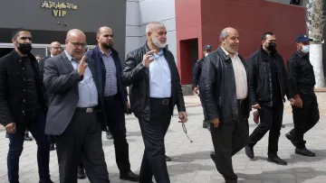 L'image montre un groupe de plusieurs hommes marchant ensemble à l'extérieur. Ils semblent se déplacer avec assurance et saluent peut-être des personnes présentes. Les hommes portent des vêtements variés, allant de vestes en cuir à des chemises et des pantalons sombres. L'environnement autour d'eux semble être un espace public, peut-être à proximité d'un bâtiment. Certains membres du groupe portent des masques. L'atmosphère paraît informelle mais organisée.