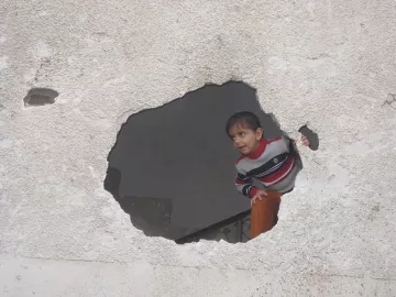 L'image montre un enfant qui regarde à travers un trou dans un mur. Le mur est en mauvais état, avec une surface rugueuse et des fissures visibles. L'enfant porte un pull rayé et semble curieux, se penchant légèrement pour observer ce qui se trouve de l'autre côté du mur. La scène évoque une atmosphère d'exploration et d'intrigue.