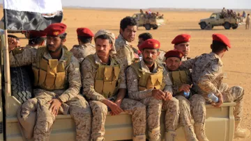 تظهر في الصورة مجموعة من الجنود المرتدين زيًا عسكريًا، يجلسون على مؤخرة مركبة عسكرية في منطقة صحراوية. يرتدي بعض الجنود قبعات حمراء، بينما يظهر العلم اليمني في الخلف. يبدو الجنود في وضعية مريحة، مع تواجد مركبات أخرى في الخلفية. تعكس الصورة أجواء عسكرية في بيئة قاحلة.