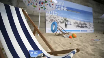 The image features a beach scene with a wooden deck chair adorned with blue and white stripes, set against a backdrop of a sandy area. In front of the chair are brochures for Tel Aviv, including one titled "Tel Aviv Gay Vibe" and another labeled "Smart City Map." The background includes a large poster depicting a sunny beach with umbrellas and people relaxing, suggesting a vibrant, beachside atmosphere.