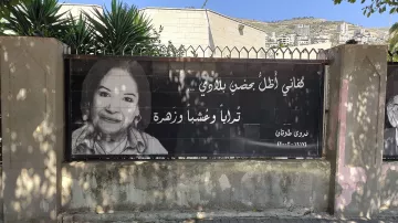 L'immagine mostra un grande striscione che riporta una fotografia di una donna sorridente, accompagnata da un testo in arabo. Il contesto sembra essere un'area urbana, con edifici sullo sfondo e piante verdi nelle vicinanze. L'atmosfera è accogliente e il messaggio sullo striscione appare positivo e invitante.
