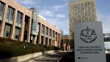 L'image montre le bâtiment de la Cour de justice des communautés européennes, situé à Luxembourg. On peut y voir une architecture moderne avec des éléments en verre et en pierre. À l'avant-plan, il y a un panneau indiquant le nom de la cour, accompagné de symboles de la justice. L'ensemble est entouré d'un aménagement paysager, avec des arbustes et un chemin qui mène à l'entrée. Le ciel est partiellement nuageux, ajoutant une ambiance calme à la scène.