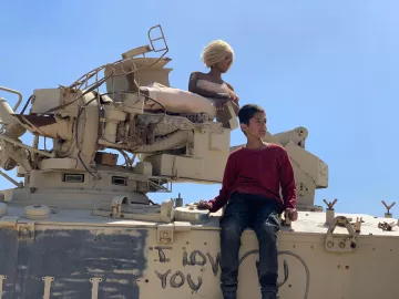 L'image montre un garçon assis sur un véhicule militaire. À côté de lui, il y a une poupée aux cheveux blonds, perchée sur le véhicule. Le fond est clair, avec un ciel bleu. Sur le véhicule, on peut voir des inscriptions, dont "I LOVE YOU" en lettres. L'ensemble de la scène dégage une atmosphere contrastée entre l'innocence de l'enfant et le décor militaire.