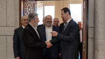 L'image montre un groupe d'hommes en train de se rencontrer dans un intérieur. Plusieurs d'entre eux sourient et semblent engagés dans une conversation amicale. L'un des hommes, au premier plan, serre la main d'un autre, tandis que les autres les observent. L'environnement semble formel, avec des éléments décoratifs visibles, comme une porte ornée et des fenêtres laissant entrer la lumière.