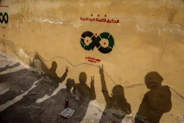 L'image montre un mur jaune où sont peints des symboles associés à un mouvement ou un parti, avec des étoiles et un motif en forme d'infini. Au premier plan, on peut voir des ombres de personnes levant les mains en signe de victoire, ce qui évoque un message de espoir et de résistance. L'atmosphère semble chargée d'émotion, probablement liée à un contexte social ou politique.