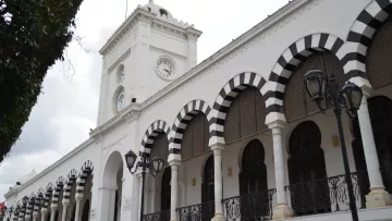 L'image montre un bâtiment architectural blanc avec des arcades noires et blanches. Il y a une horloge sur la façade et un drapeau tunisien flottant au sommet. Le ciel est nuageux, ce qui donne une ambiance légèrement sombre. Des lampadaires sont visibles le long de la structure, ajoutant une touche d'élégance à l'ensemble.