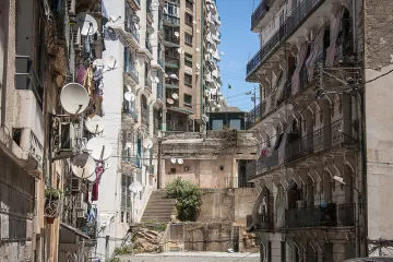 L'image montre une vue urbaine d'une rue avec des bâtiments en pierre et des murs en béton. On observe un mélange d'architecture ancienne et moderne, avec des balcons ornés de fer forgé. De nombreux paraboles satellites sont installées sur les façades des bâtiments, tandis que des linges pendus à l'extérieur ajoutent une touche de vie quotidienne. Les escaliers menant à d'autres niveaux sont visibles, et le ciel est clair et ensoleillé, donnant une atmosphère vive à la scène.