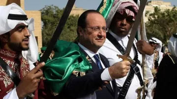 L'image montre un homme au centre, portant une veste sombre, accompagné de deux personnes de chaque côté. Ces dernières sont vêtues d'habits traditionnels arabes et tiennent des épées. L'arrière-plan montre un ciel clair et un environnement désertique ou historique, suggérant une ambiance festive ou cérémonielle. L'expression de l'homme central semble joyeuse et accueillante, indiquant qu'il participe à une célébration ou un événement culturel.