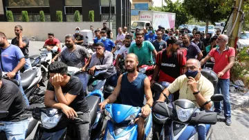 در این تصویر، گروهی از مردان در حال انتظار بر روی موتور سیکلت‌های خود هستند. برخی از آنها در حالت بی‌حوصله به نظر می‌رسند و دیگران مشغول صحبت یا چک کردن گوشی‌های همراه خود هستند. وضعیت به نظر شلوغ و شلوغی در اطراف وجود دارد. اکثر مردان تی‌شرت‌های متنوعی پوشیده‌اند و فضای عمومی در پس‌زمینه به وضوح دیده می‌شود.
