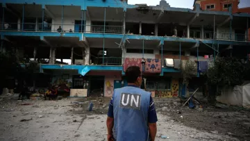تظهر في الصورة شخص يرتدي سترة تحمل شعار الأمم المتحدة (UN)، يقف في مكان يبدو متضرراً بشدة. خلفه توجد عمارة متضررة، مع آثار دمار واضحة على الجدران، حيث تتواجد شرفات وبعض النوافذ المكسورة. الأرض مغطاة بالأنقاض والحطام، مما يدل على أن المكان تعرض لأحداث قد تكون كارثية أو نزاع. الجو العام يوحي بالحزن والخراب.
