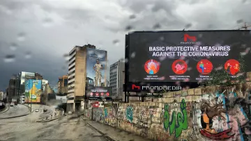 La imagen muestra una calle en un entorno urbano, con un ambiente lluvioso. Hay un letrero digital grande que presenta medidas de protección básicas contra el coronavirus. En el fondo, se pueden ver edificios y murales coloridos en las paredes, lo que añade un contraste visual a la escena. El cielo está nublado, lo que sugiere un clima gris y lluvioso.