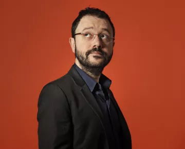 L'image montre un homme vêtu d'un costume noir sur une chemise bleu foncé, regardant légèrement sur le côté. Il a des lunettes, une barbe et un léger sourire. L'arrière-plan est orange, ce qui crée un contraste vif et dynamique avec sa tenue sombre. L'expression de son visage semble pensif ou intrigué.