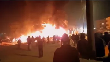 L'image montre une scène nocturne où un bâtiment est enflammé, créant une grande colonne de feu et de fumée. Des personnes se tiennent au premier plan, observant la situation. On peut noter une ambiance de tension, avec des lumières des bâtiments environnants qui ajoutent à l'atmosphère dramatique. Les silhouettes des personnes sont découpées contre la lumière des flammes.