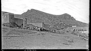 L'image présente un paysage désertique avec des montagnes en arrière-plan. À gauche, on aperçoit des bâtiments en pierre, probablement des fortifications ou des maisons, qui semblent très anciens. Au premier plan, un groupe de personnes est visible, accompagné de plusieurs animaux de charge, comme des chameaux ou des ânes. Le ciel est clair, montrant une atmosphère paisible, et le terrain est aride, typique des régions montagneuses désertiques. L'ensemble dégage une impression d'histoire et de culture.