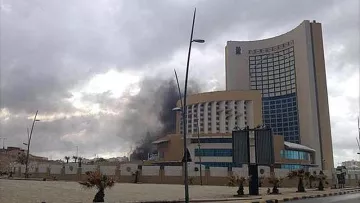 L'image montre un bâtiment moderne de grande taille, probablement un hôtel ou un immeuble de bureaux, situé dans une zone urbaine. À l'arrière-plan, on peut voir de la fumée noire s'élever dans le ciel, ce qui suggère qu'il y a eu un incendie ou une explosion. Le temps semble nuageux, ajoutant une atmosphère sombre à la scène. Des lampadaires bordent la route, et il y a des espaces verts et des plantations autour du bâtiment.