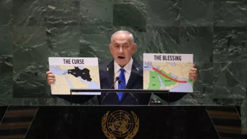 L'image montre un homme debout derrière un pupitre, s'exprimant lors d'un discours. Il tient deux panneaux, l'un portant l'inscription "THE CURSE" (la malédiction) et l'autre "THE BLESSING" (la bénédiction). Sur ces panneaux, il y a des cartes représentant des régions du Moyen-Orient, probablement pour illustrer un point sur des questions géopolitiques. Le décor est formel, semblant indiquer une réunion internationale, comme celle des Nations Unies. L'homme porte une cravate et une veste, se tenant de manière convaincante.