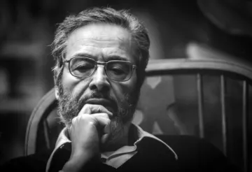 The image features a man with a thoughtful expression, sitting in a wooden chair. He has a beard and glasses, and he appears to be deep in contemplation. The lighting is soft, creating a moody atmosphere, and the background is blurred, focusing attention on his contemplative demeanor. The photograph has a classic black-and-white aesthetic, emphasizing the seriousness of the moment.