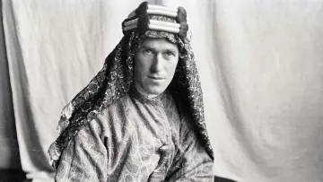L'image montre un homme portant un costume traditionnel. Il est vêtu d'une tunique ornée et d'un headdress décoratif. Son regard est sérieux et il tient un livre dans ses mains. L'arrière-plan est sobre, ce qui met en valeur son apparence et son habillement. La photo semble être ancienne, ce qui lui donne un aspect historique.
