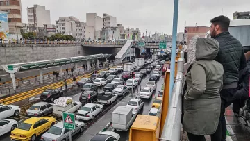 L'image montre une scène de circulation dense dans une ville. On peut voir de nombreuses voitures de différentes marques immobilisées dans un embouteillage. À l'arrière-plan, on distingue des bâtiments urbains et une route à plusieurs niveaux. Il y a des panneaux de signalisation visibles, et deux personnes se tiennent sur un balcon ou une balustrade, observant la circulation en contrebas. Le ciel est nuageux, ce qui suggère un temps gris et éventuellement pluvieux.