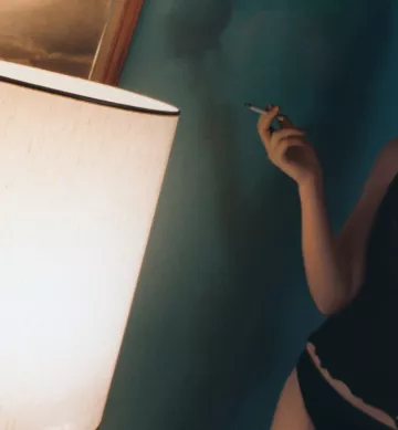 L'image montre un intérieur avec une lumière douce émise par une lampe. Une personne, située à gauche, tient un objet à la main qui semble être une cigarette. La décoration murale est d'une teinte douce, créant une atmosphère intime et relaxante. La lumière de la lampe joue sur les surfaces, ajoutant une ambiance chaleureuse à la scène.