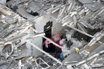 L'image montre des ruines d'un bâtiment, avec des débris de toit et des murs effondrés. Deux personnes sont visibles à l'intérieur des décombres, semblant chercher ou s'entraider dans cet environnement chaotique.