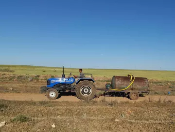 L'image montre un homme conduisant un tracteur bleu sur une route de terre. Il tire une remorque avec un grand réservoir métallique. À l'arrière-plan, on peut voir des champs verts sous un ciel dégagé. La scène évoque une ambiance rurale et agricole.
