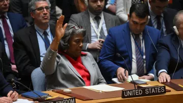 L'immagine mostra un incontro del Consiglio di Sicurezza delle Nazioni Unite. Una donna, che sta al centro della scena, alza la mano, apparentemente per esprimere un'opinione o richiedere la parola. Indossa un blazer grigio con una camicia rossa e sembra essere in fase di comunicazione. Attorno a lei, ci sono diversi rappresentanti di altri paesi, tra cui stati come Regno Unito e altri, seduti con atteggiamenti attenti. L'atmosfera sembra seria e formale, tipica di una riunione diplomatica.