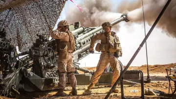 L'image montre deux soldats en uniforme militaire, probablement dans un environnement désertique. Ils sont proches d'une grande pièce d'artillerie qui est en train de tirer, créant un nuage de fumée. L'un des soldats semble gérer l'équipement tandis que l'autre se tient debout, surveillant la situation. Un camouflage est utilisé pour dissimuler l'équipement militaire, et le paysage environnant est aride avec un ciel dégagé. L'atmosphère semble tendue et concentrée, rappelant des opérations militaires.