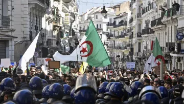 En la imagen se puede ver una multitud reunida en una calle, con varias personas sosteniendo banderas argelinas y carteles. El ambiente parece ser de una manifestación o protesta, ya que hay un grupo de policías en primer plano, equipados con cascos y protecciones, observando la situación. Los edificios a ambos lados de la calle son visibles, creando un contexto urbano y de actividad social.