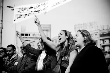 L'image montre un groupe de personnes participant à une manifestation. On peut voir des femmes au premier plan levant le poing et chantant, exprimant des émotions fortes et de la détermination. L'ambiance semble dynamique et engageante. En arrière-plan, des banderoles apparaissent, suggérant un message ou une demande collective. La photo est en noir et blanc, ce qui accentue l'intensité et l'impact visuel de la scène.