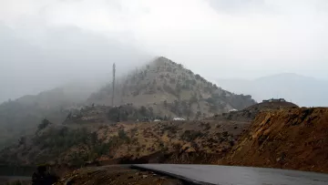 L'image montre un paysage montagneux avec des collines voilées par le brouillard. En arrière-plan, une structure de télécommunication est visible au sommet d'une colline. La route qui serpente est goudronnée, et le terrain environnant présente des nuances de terre brune et de verdure. L'atmosphère semble sombre et mystérieuse, suggérant une journée nuageuse et humide.