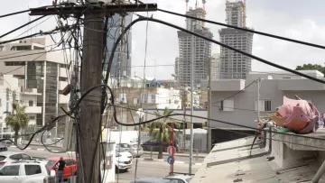 L'image montre une vue urbaine où plusieurs poteaux électriques sont entremêlés de fils. À l'arrière-plan, on peut apercevoir deux grands immeubles en construction. Au premier plan, il y a des bâtiments plus petits et des véhicules stationnés. On peut également voir des palmiers, ce qui indique un environnement peut-être chaud ou méditerranéen. L'ensemble donne une impression de ville en développement, avec un mélange de structures anciennes et modernes.