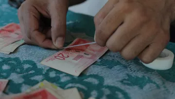 L'image montre des mains qui manipulent des billets de banque. Une personne semble être en train de recoller ou d'ajuster un billet de 20 unités. On peut voir un surface en tissu avec des motifs, ainsi que d'autres billets éparpillés autour. La situation donne une impression de travail minutieux avec une attention particulière aux détails.